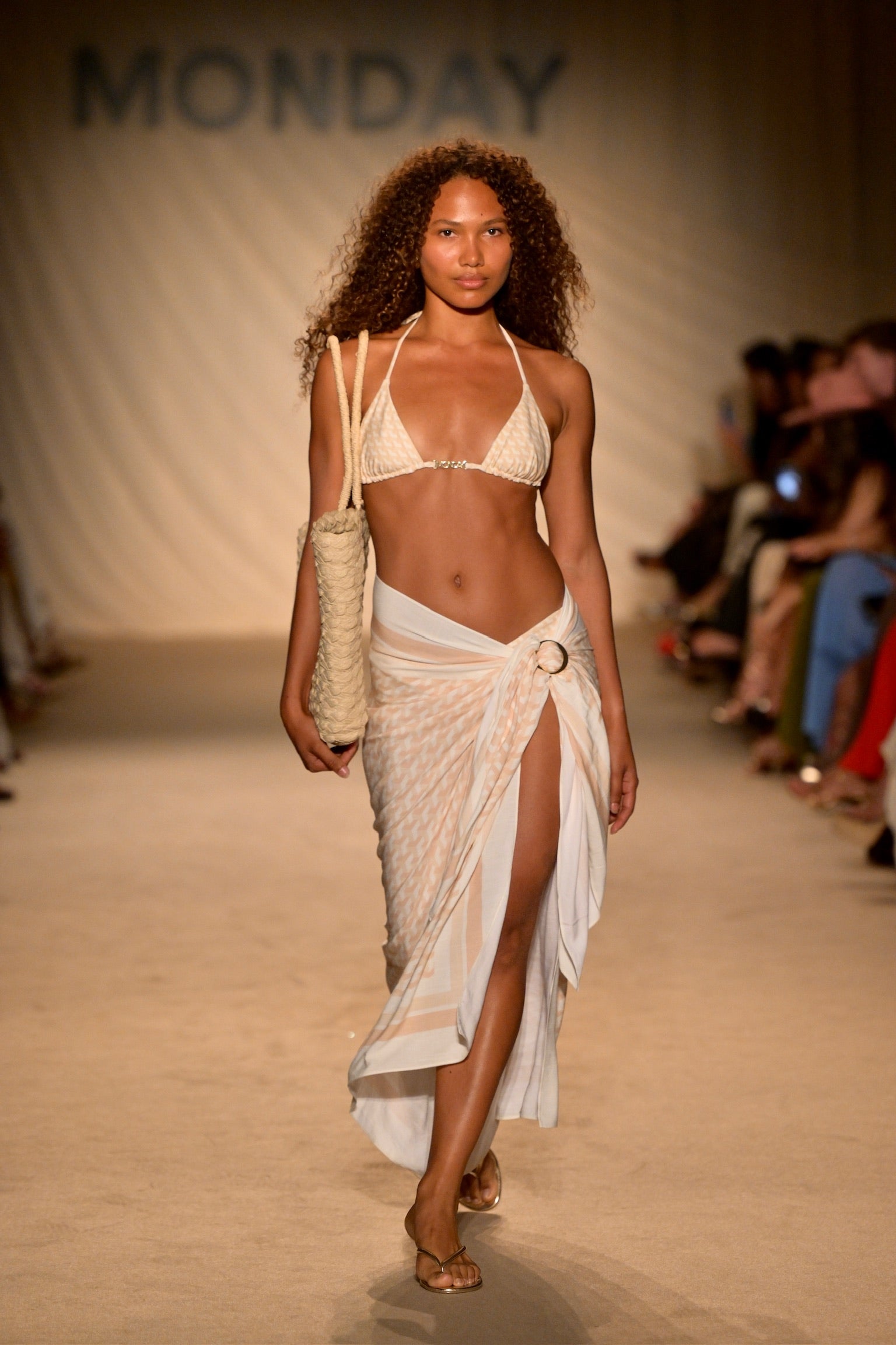 Model in cream bikini and sheer wrap skirt carrying a woven tote on runway; backdrop text reads KOH SWIM.