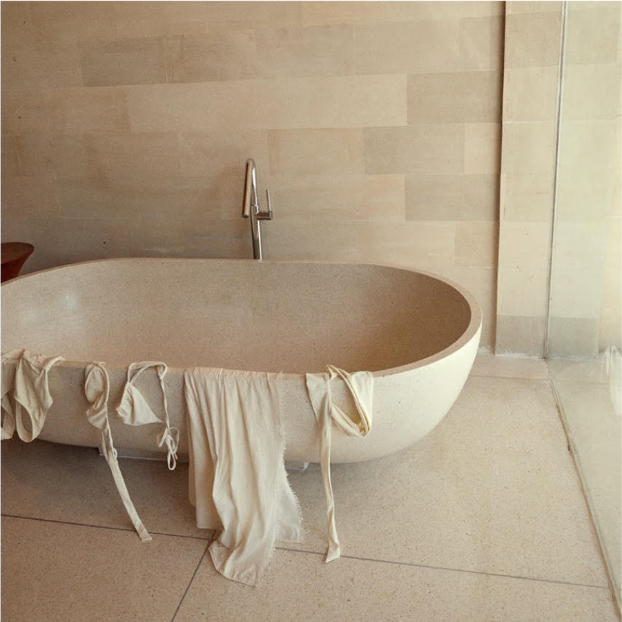 Freestanding beige stone bathtub with cream towels draped over the edge, wall-mounted faucet and tiled wall.