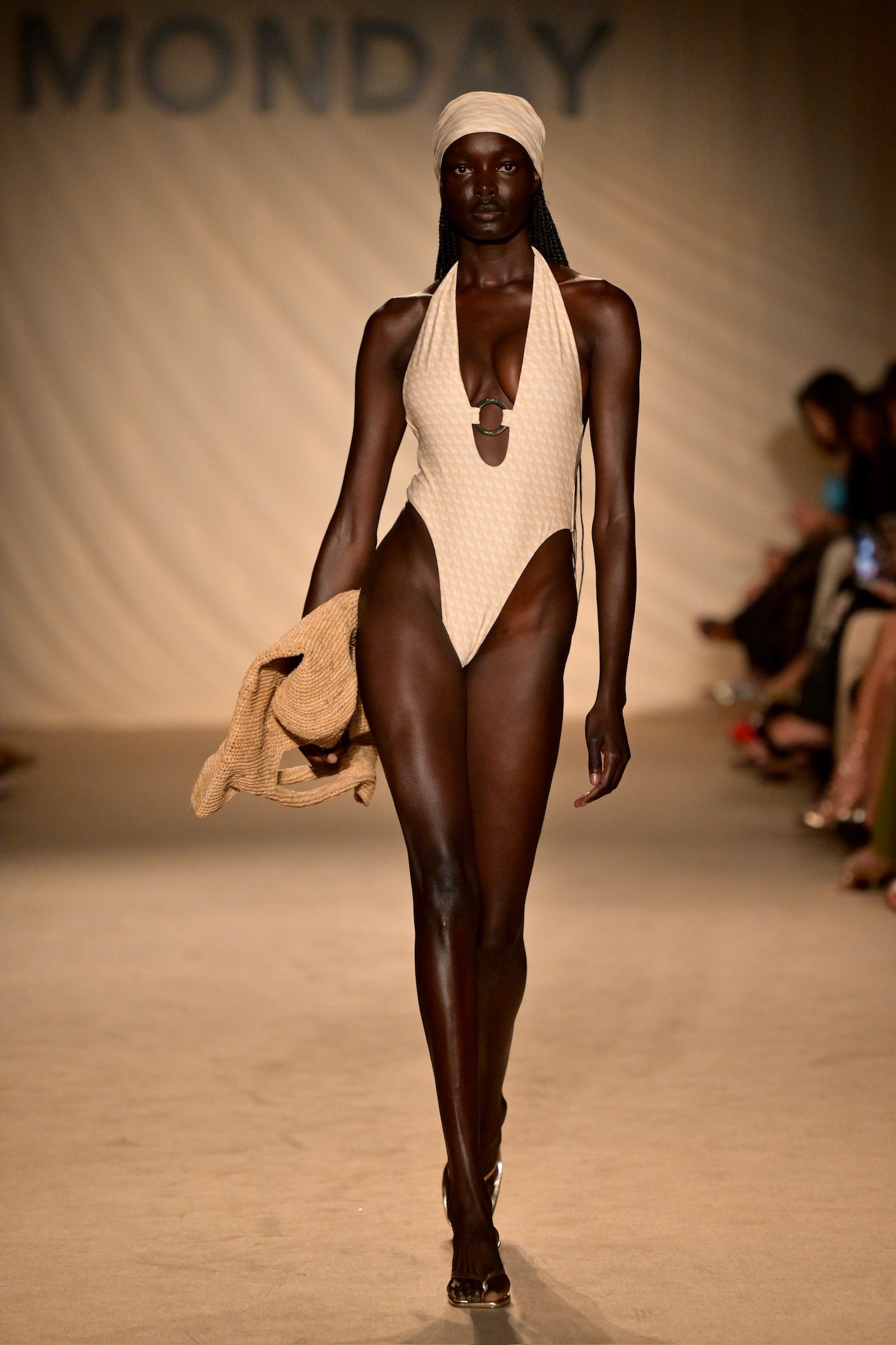 Dark-skinned runway model in beige high-cut swimsuit and headscarf holding woven jacket; KOH SWIM on backdrop.